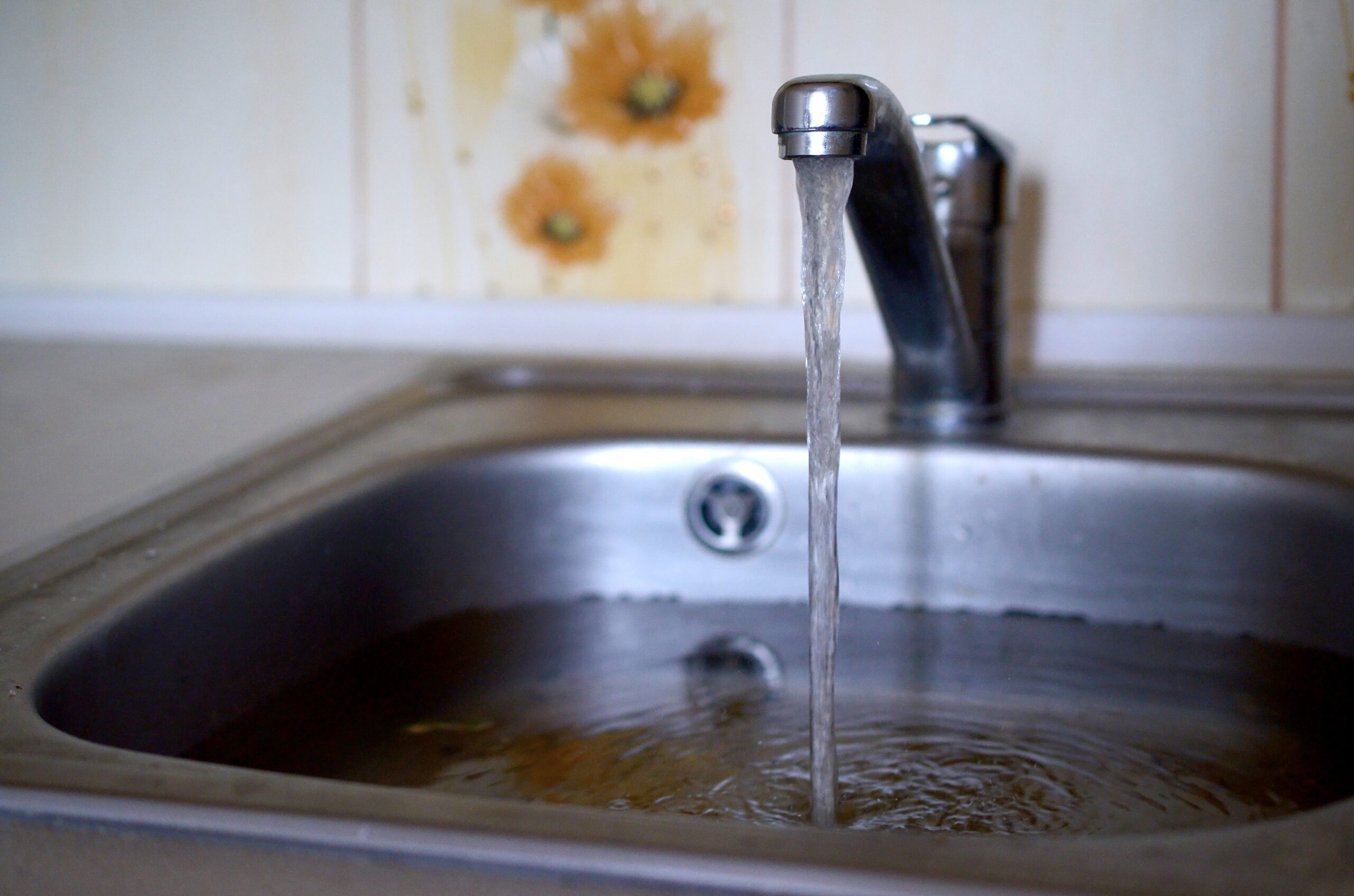 %Title% | Aaa Service Network An AAA Service Network technician tests a kitchen faucet as water flows into a stainless steel sink, backed by floral-patterned tiles.