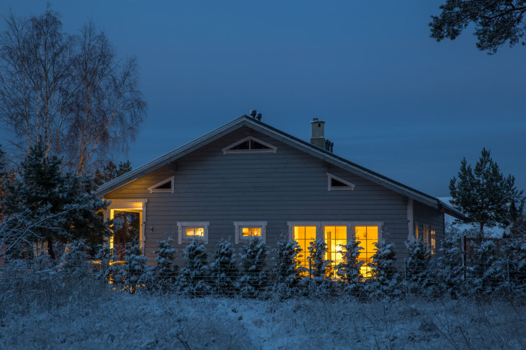 A cozy, glowing home nestled in snowy trees at dusk stays warm, powered by a Whole Home Generator from AAA Service Network.