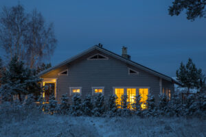 A wooden house with glowing yellow windows stands in a snowy landscape surrounded by trees in the evening, its warmth ensured by a Whole Home Generator.