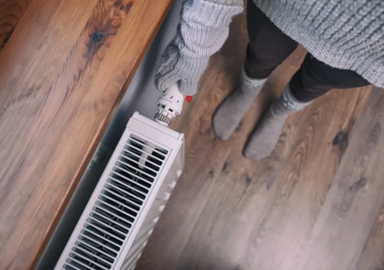 %Imagetitle% | Aaa Service Network Person wearing gray sweater, gloves, and socks adjusts a radiator thermostat on a wooden floor next to a wooden bench or windowsill.