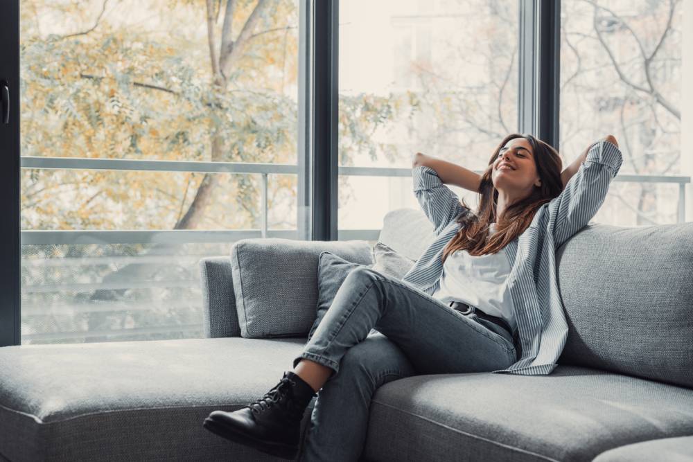 A AAA Service Network customer relaxes on a gray sofa near large windows, while our air purifier ensures fresh, clean indoor air.