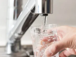 A hand holds a clear glass under a metal faucet as water pours into the glass, with bubbles forming inside—demonstrating the clarity often achieved with a water softener.