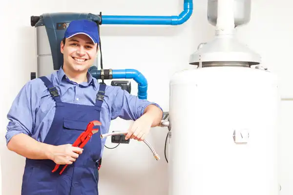 An AAA Service Network plumber in uniform smiles by a water heater, holding pipe wrenches and a flexible tube, ready to help.