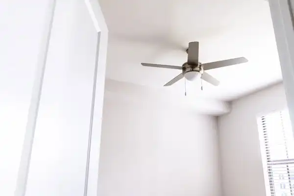 A white ceiling fan with a light fixture is installed in a bright, tidy room with a window and blinds by AAA Service Network.