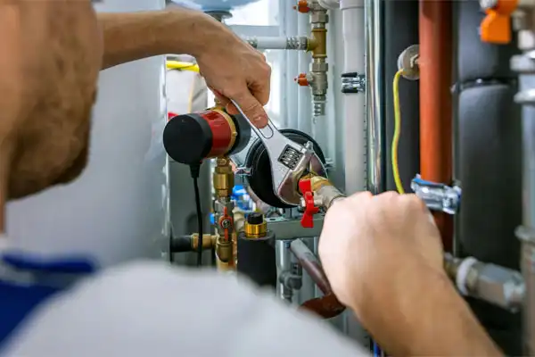 An AAA Service Network technician adjusts plumbing connections with a wrench on pipes and valves in a utility room.