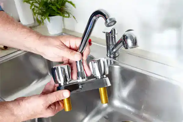 An AAA Service Network technician prepares to install a new kitchen faucet, holding it above a gleaming stainless steel sink.