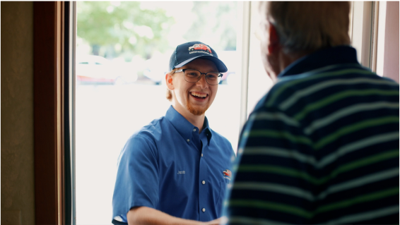 A delivery person in a blue uniform and cap smiles while greeting a man at a doorway.